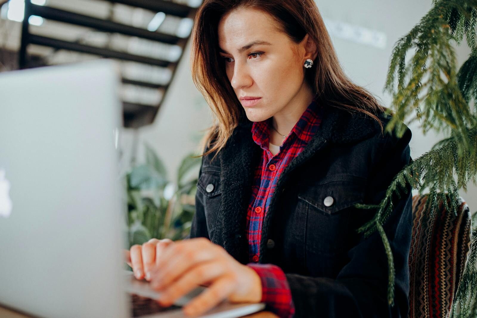 A serious woman in a plaid shirt works intently on her laptop indoors.